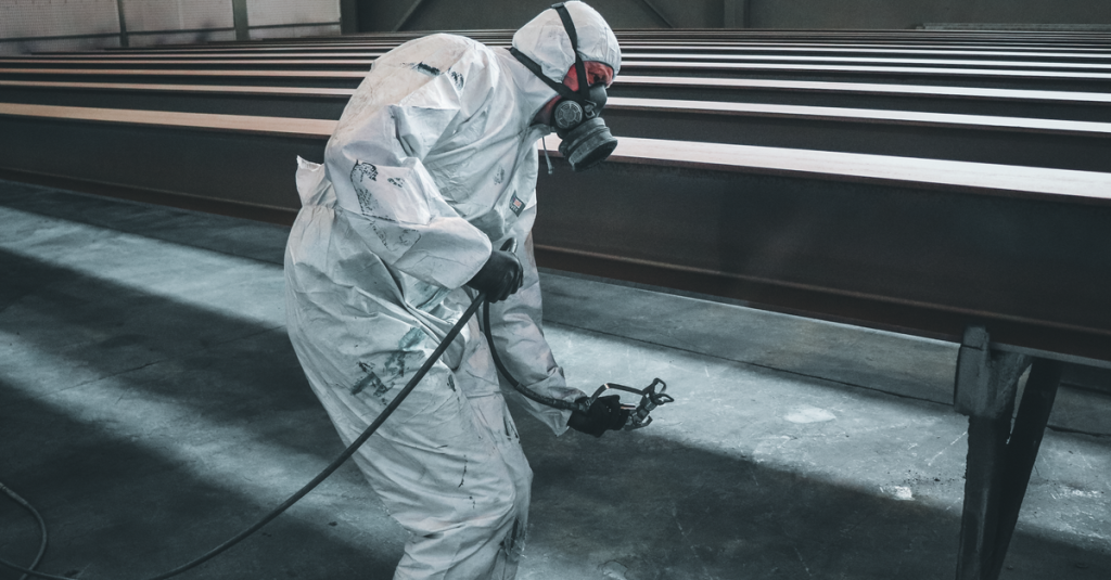 Worker in protective gear spray painting metal beams inside a factory setting.