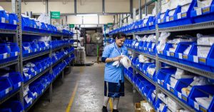 A woman organizing inventory in a warehouse aisle, surrounded by shelves and blue bins.