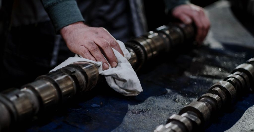 Close-up of a mechanic cleaning a camshaft with a cloth in an industrial setting.