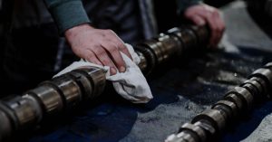 Close-up of a mechanic cleaning a camshaft with a cloth in an industrial setting.