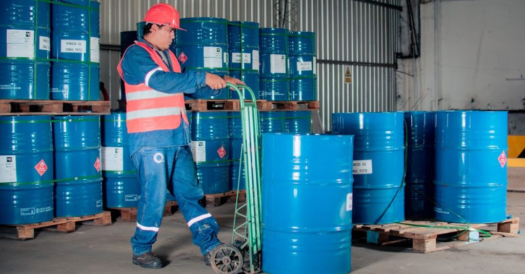 Industrial worker with a hand truck moving blue barrels in a warehouse setting.