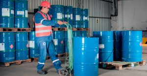 Industrial worker with a hand truck moving blue barrels in a warehouse setting.
