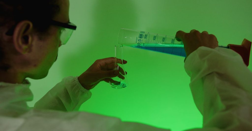 Scientist carefully pouring blue liquid into a test tube under green light.